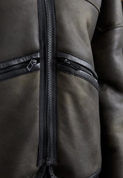 Dunkelgrüne Lederjacke mit asymmetrischen Reißverschlüssen, strukturiertem Finish, schwarzen Akzenten und zwei Reißverschlussfronttaschen.