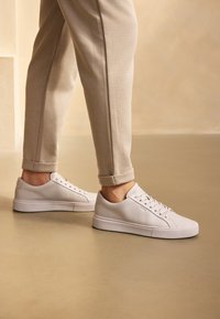 Zapatillas de deporte de cuero blanco con una textura suave, con un diseño de cordones y una suela de goma gruesa. Combinadas con pantalones beige enrollados.