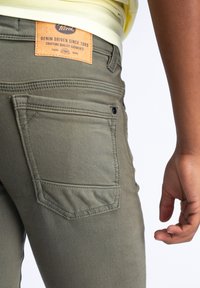 Olivengrønne denimbukser med et lædermærke, der læser "Petrol" på bagsiden af taljen; de har en enkelt lomme og synlige sy-detaljer.
