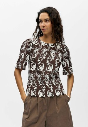 Femme aux cheveux ondulés foncés portant un haut marron à manches courtes avec une broderie florale blanche et un pantalon marron plissé ample, mains dans les poches.
