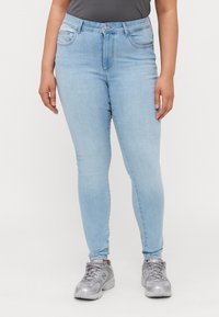 Jeans ajustados de color azul claro hechos de mezclilla, con cintura media, cinco bolsillos y pasadores para cinturón. Combinados con zapatillas deportivas grises.