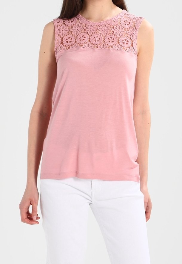 Haut rose sans manches avec empiècement en dentelle florale porté avec un pantalon blanc, montrant le haut du corps de face jusqu'aux hanches.