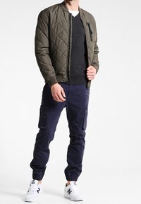 Veste bomber en olive matelassée sur un pull noir, associée à un pantalon cargo marine et des baskets blanches. La veste possède une poche zippée.