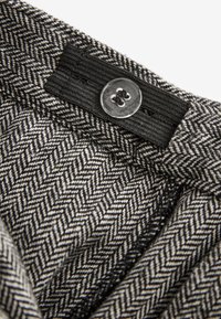 Textília s kostkovaným vzorom houndstooth v čierno-bielej farbe, s čiernym pásom a okrúhlym priehľadným gombíkom na zapínanie.