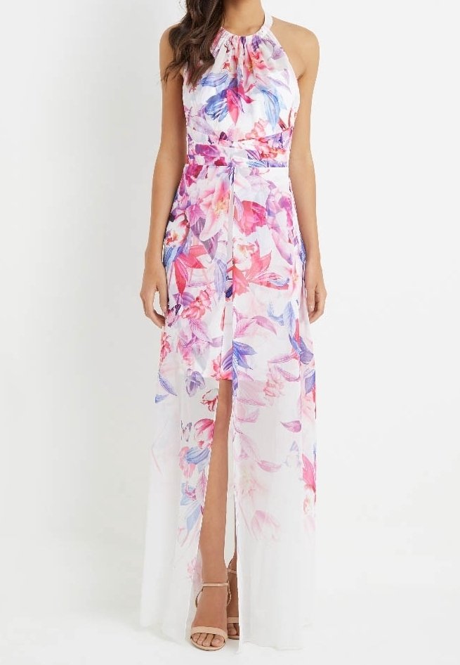 Femme portant une robe maxi blanche sans manches avec imprimé floral rose et bleu et une fente à l'avant, associée à des talons beiges à bride à la cheville.