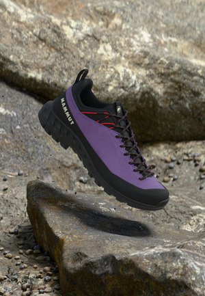 Lila és fekete Mammut túracipő piros fűzőkkel, amely egy sziklás szabadtéri terület fölött lebeg.