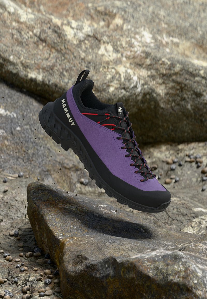 Μωβ και μαύρο ορειβατικό παπούτσι Mammut με κόκκινα κορδόνια που αιωρείται πάνω από βραχώδη εξωτερική περιοχή.