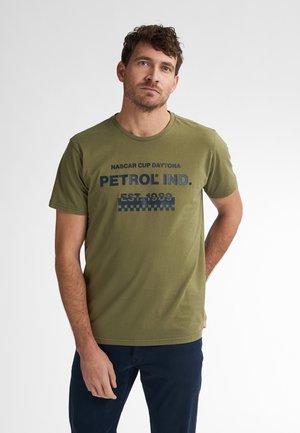 T-shirt en coton vert olive avec un imprimé graphique bleu foncé portant la mention "PETROL IND." et un motif à carreaux en bas. Manches courtes, col rond.