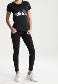 Camiseta negra de Adidas con logo blanco, combinada con leggings negros y zapatillas deportivas blancas. Diseño simple y ajustado con cuello redondo.