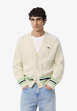 Cardigan en tricot torsadé crème avec des manches longues, fermeture à boutons et rayures vertes et bleues marine à l'ourlet. Présente un petit logo sur la poitrine.