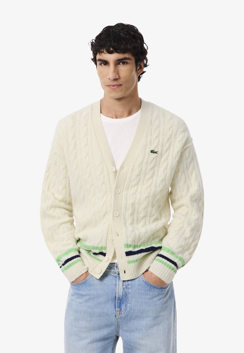Cardigan a maglia a trecce color crema con maniche lunghe, chiusura con bottoni e strisce verde e blu navy all'orlo. Presenta un piccolo logo sul petto.