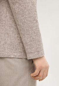 Primo piano della mano di una persona che riposa accanto alla manica di una giacca beige testurizzata e a un paio di pantaloni beige chiaro, con una cucitura della tasca visibile.