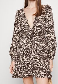 Nainen, joka on pukeutunut pitkähiharaiseen leopardikuosiseksi mekkoon, jossa on syvä V-mallinen kaula-aukko ja edessä solmittava yksityiskohta, röyhelöinen helma ja vaalean beige tausta.