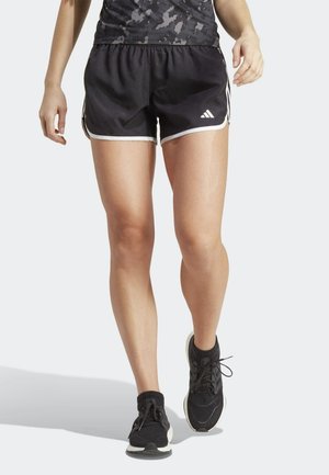 Kobieta nosząca czarne krótkie spodenki do biegania Adidas, czarne buty sportowe i bluzkę z wzorem moro, idąca naprzód na jednolitym tle.