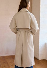 Beige trench coat med brede ærmer, bælte i taljen og rygslids. Fremstillet af glat materiale og designet til en afslappet pasform.