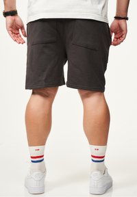 Dunkelgraue Baumwollshorts mit zwei Gesäßtaschen. Das Modell trägt weiße Sneaker und Socken mit roten und blauen Streifen am oberen Ende.