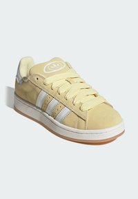 Zapatillas de ante amarillo con rayas blancas, logo redondo en la lengüeta, cordones gruesos y suela de goma. Textura suave y diseño de perfil bajo.