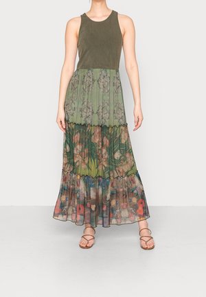 Frau trägt ein ärmelloses, olivgrünes, figurbetontes Oberteil mit langem, gestuftem, geblümtem Maxirock und braunen Riemchensandalen.