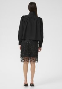 Blouse noire courte avec des manches volumineuses, associée à une jupe noire transparente ornée de perles intricatement travaillées et de motifs floraux.