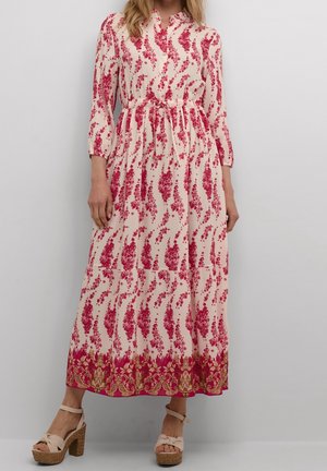 Kobieta ubrana w długą kremową suknię z czerwonymi kwiatowymi wzorami i ozdobną czerwonozłotą bordiurą, w towarzystwie beżowych sandałów na platformie.