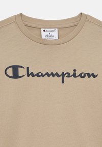 Μπεζ βαμβακερό μπλουζάκι με κοντά μανίκια, στρογγυλή λαιμόκοψη και μεγάλο σκούρο μπλε λογότυπο "Champion" τυπωμένο στο εμπρός μέρος.