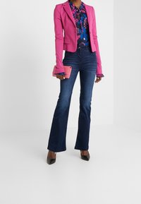Blazer ajustado rosa sobre una blusa floral, jeans oscuros de campana y tacones negros de punta. Sostiene un pequeño clutch rosa.