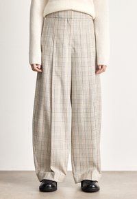 Calças de perna larga com padrão xadrez bege claro, apresentando pregas frontais proeminentes e textura suave, combinadas com sapatos pretos.