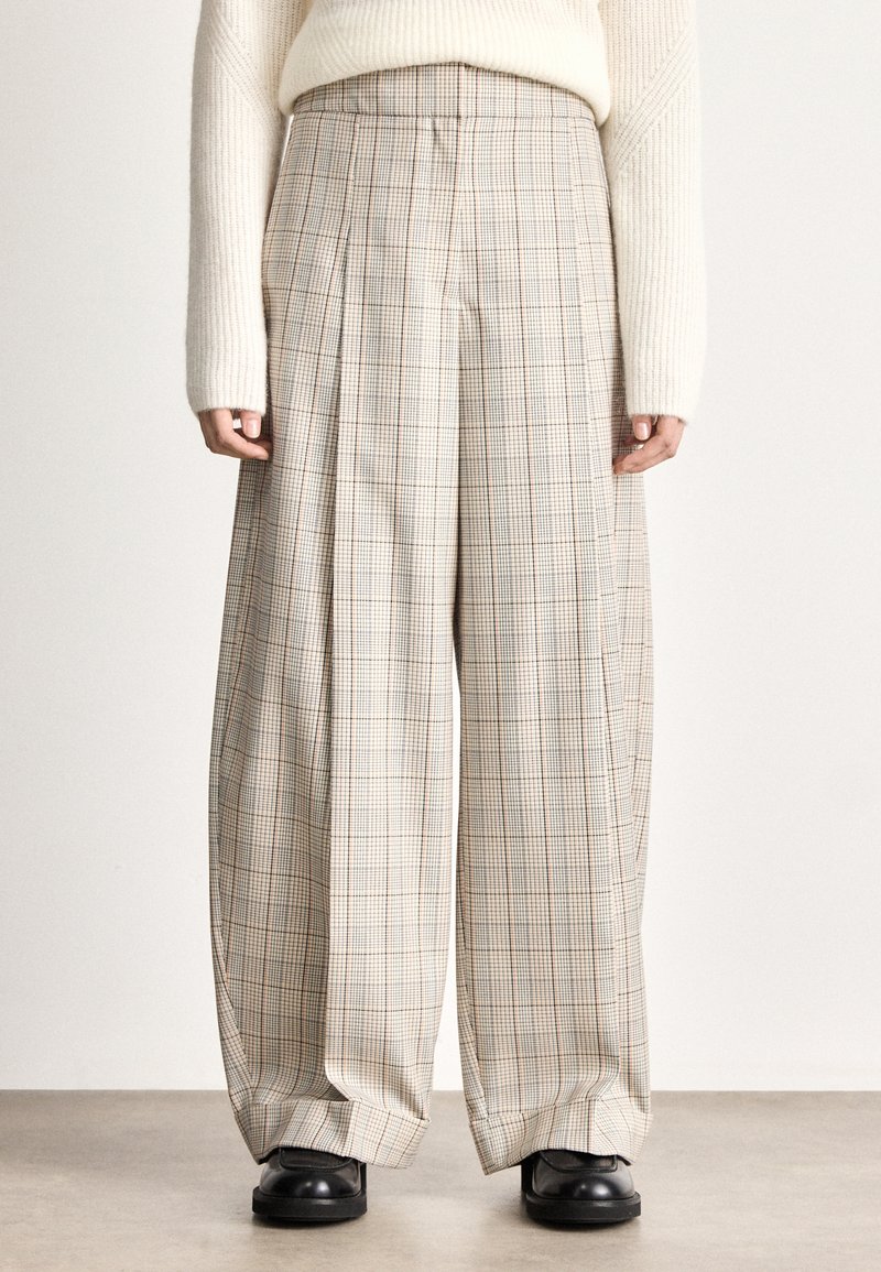Calças de perna larga com padrão xadrez bege claro, apresentando pregas frontais proeminentes e textura suave, combinadas com sapatos pretos.