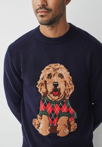 Granatowy sweter z teksturowanym, dzianinowym wzorem, na którym znajduje się brązowy pies w w kratkę czerwono-zielonym swetrze.