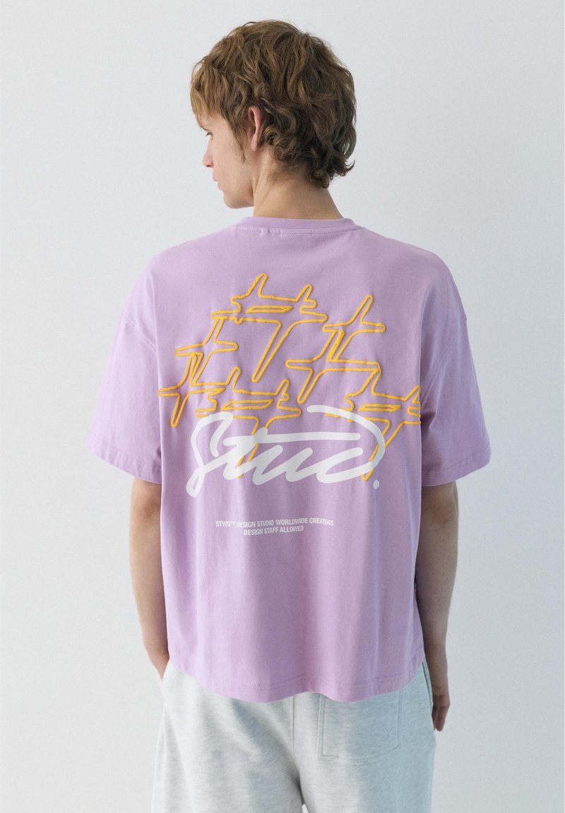 Jeune homme portant un T-shirt lavande ample avec des motifs d'avions jaunes et un texte blanc en script au dos, associé à un pantalon gris clair.