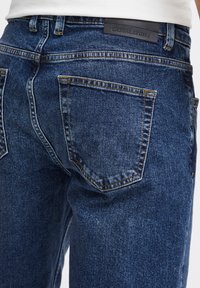 Tamno plavi traperice od jeansa s teksturiranim završetkom, s dva stražnja džepa i vidljivim šavovima. Crna oznaka nosi natpis "CASUAL FRIDAY."