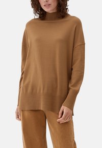 Bruine gebreide trui met een hoge hals en doorgezakte schouders. Voorzien van geribbelde manchetten en zoom. Zachte textuur, losse pasvorm, gecombineerd met beige broek.