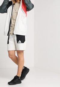 Veste pour jeune en blanc, gris et rouge avec fermeture éclair ; t-shirt beige en dessous ; short blanc avec des accents noirs ; baskets noires.