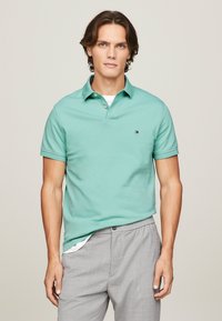 Polo de color verde claro hecho de algodón, con cuello y tres botones. Un pequeño logo está en el lado izquierdo del pecho. Combinado con pantalones grises.