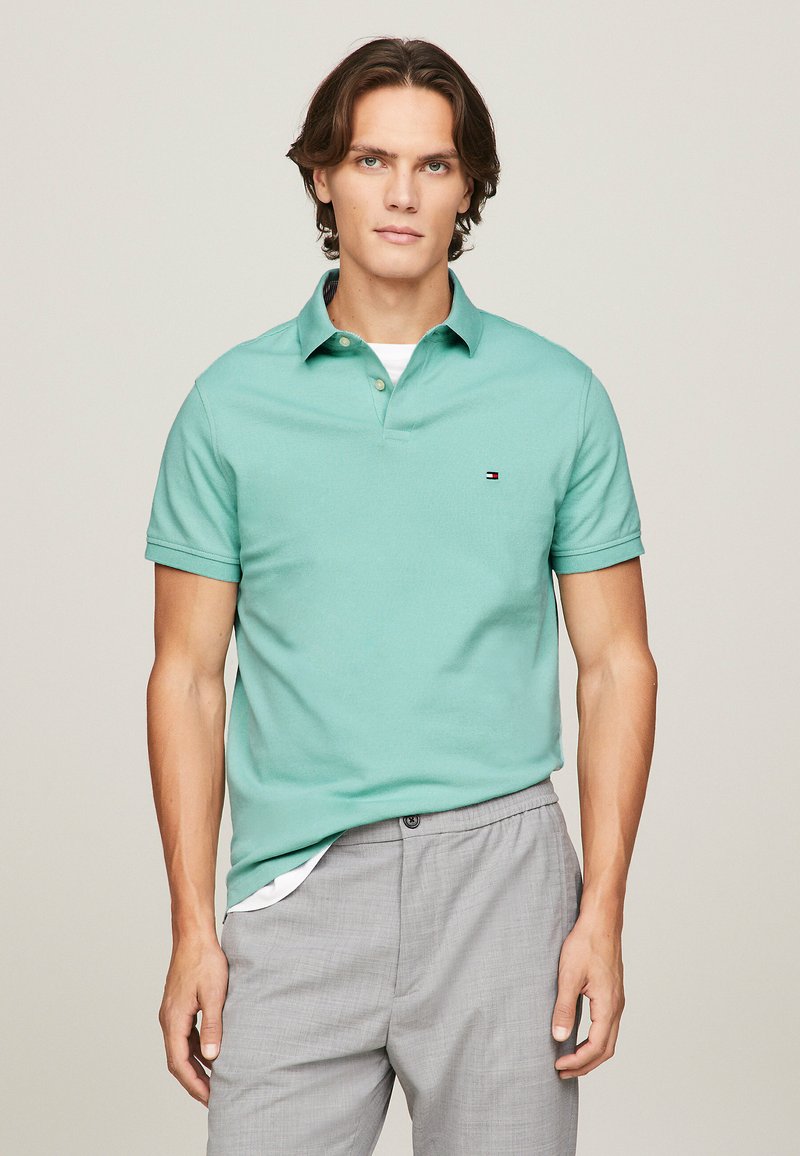 Polo de color verde claro hecho de algodón, con cuello y tres botones. Un pequeño logo está en el lado izquierdo del pecho. Combinado con pantalones grises.