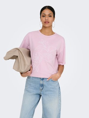 Mujer con camiseta de color rosa claro con bordados florales y vaqueros azul claro, sosteniendo una chaqueta beige, de pie frente a un fondo blanco.