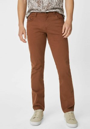 Mężczyzna w brązowych spodniach o prostych nogawkach, beżowych codziennych trampkach i wzorzystej koszuli z krótkim rękawem, stojący na jednolitym tle.