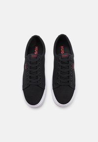 Svart kanvas sneakers med vita gummisulor, med snörning och röd "HUGO" branding på sidan och innersulan.