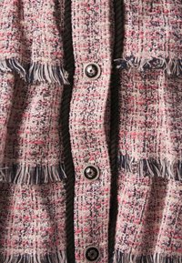 Veste en tweed tissé aux fils rose, marine et argenté ; ornée de franges et dotée de boutons noirs sur le devant pour la fermeture.