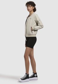 Jachetă bomber de culoare bej deschis cu fermoar frontal, manșete ribbed, buzunare laterale și accente negre. Purtați-o peste un top negru fără mâneci și pantaloni scurți. Încălțăminte sport neagră cu glezna înaltă.