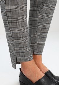 Grijze geruite broeken met zwart-witte ruiten, voorzien van zijsplitten aan de onderkant. Gecombineerd met zwarte textuur instappers.
