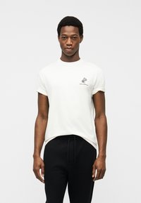 Νεαρός άνδρας όρθιος, κοιτάζοντας προς τα εμπρός, φοράει λευκό κοντομάνικο T-shirt με μικρό λογότυπο φοίνικα και μαύρο παντελόνι.