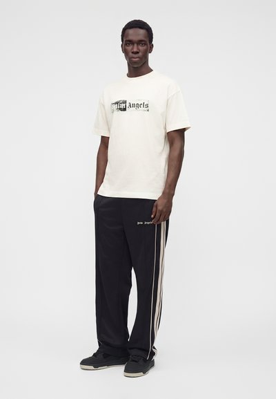 Νέος άνδρας στέκεται, φοράει ένα λευκό t-shirt Palm Angels και μαύρο παντελόνι φόρμας με λευκές ρίγες στα πλαϊνά, καθώς και μαύρα αθλητικά παπούτσια.
