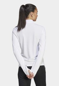 Weißes Langarm-Oberteil mit strukturiertem Muster, separatem Kragen und Daumenlöchern. Getragen mit schwarzen Hosen, von hinten betrachtet.