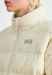 Beige puffer jacket with high collar, zip closure, and a logo in light gray. Features quilted texture and smooth, shiny material.