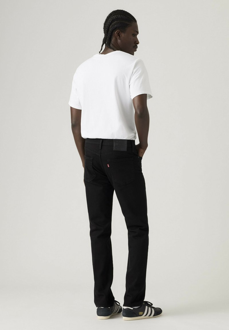 Mann mit geflochtenem Haar, der ein weißes T-Shirt, schwarze Jeans und schwarze Adidas-Sneaker trägt, steht mit den Händen in den Taschen und blickt seitlich.