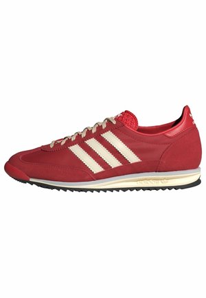 Czerwony but sportowy Adidas z białymi paskami, beżowymi sznurówkami, teksturowaną podeszwą oraz oznakowanym obcasem i środkową częścią podeszwy, pokazany z profilu z boku, skierowany w lewo.
