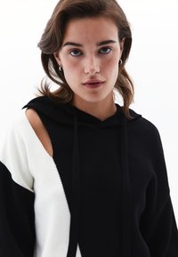 Schwarzer und weißer Hoodie mit diagonaler Design, offener Schulter, Kordeln und einem weichen, strukturierten Stoff.