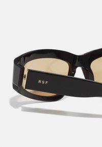 Sorte solbriller med tykke rammer, gull RSF-merkevare på armen, og tonet brune linser. Glatt plastmateriale og vinkeldesign.