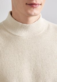 Cremefarbener Rollkragenpullover mit gerippter Textur und enganliegendem Hals. Nahaufnahme, die die Weichheit und das Muster des Stoffes hervorhebt.